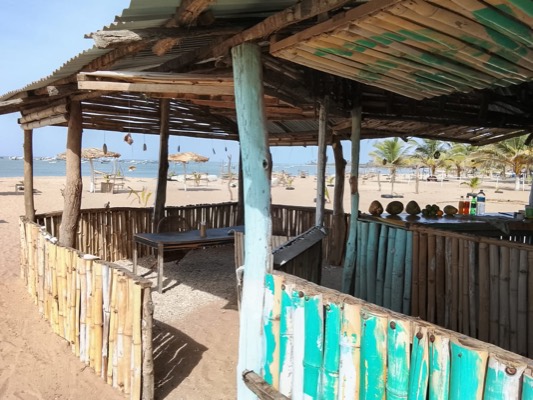 The palm-thatch bar at Coco Palace with its painted bamboo counter, looking out across the sand to the parasols and the sea.